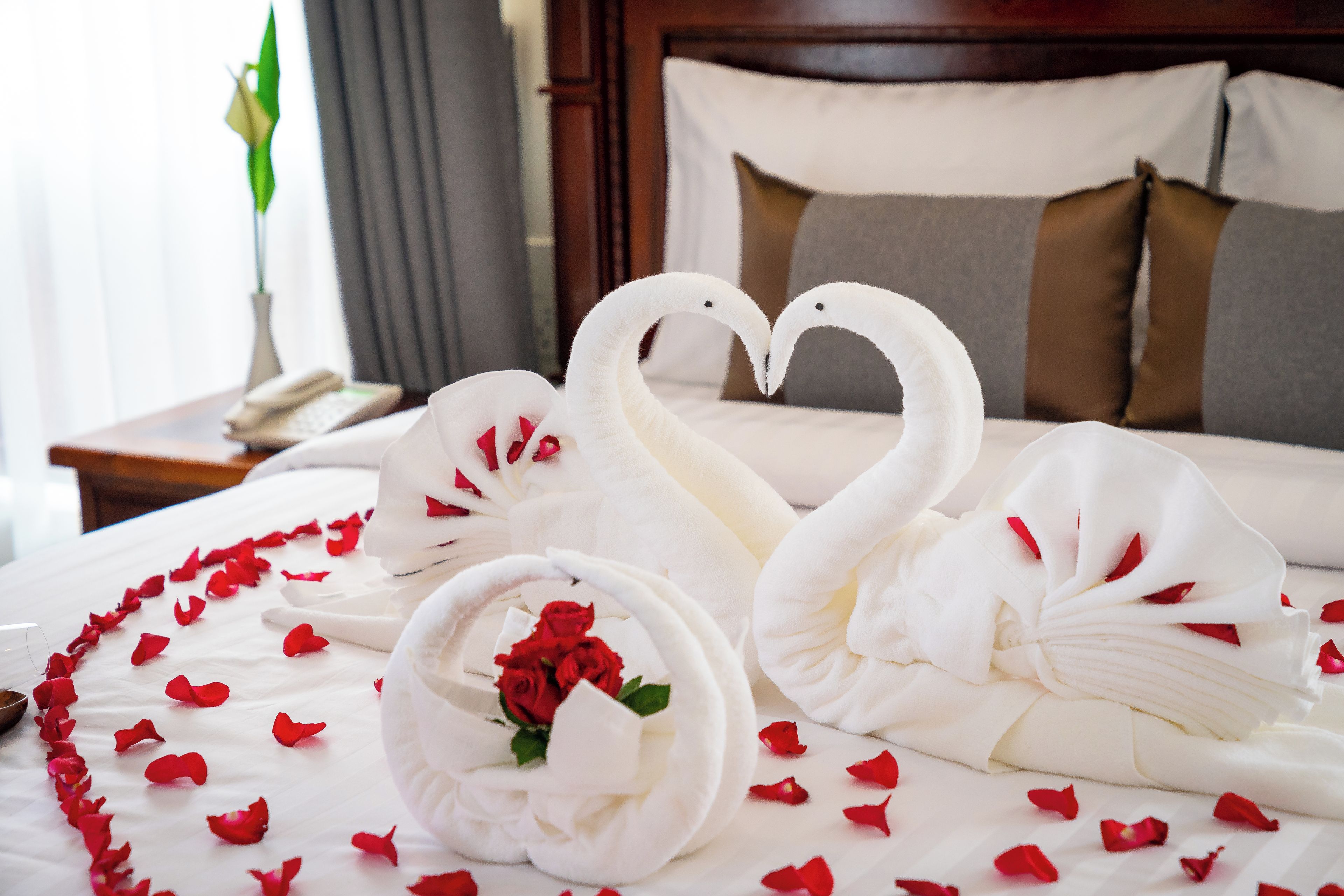 A bed with white linens, a heart-shaped arrangement of towels, and scattered rose petals.