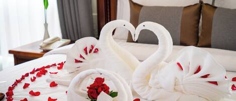 A bed with white linens, a heart-shaped arrangement of towels, and scattered rose petals.