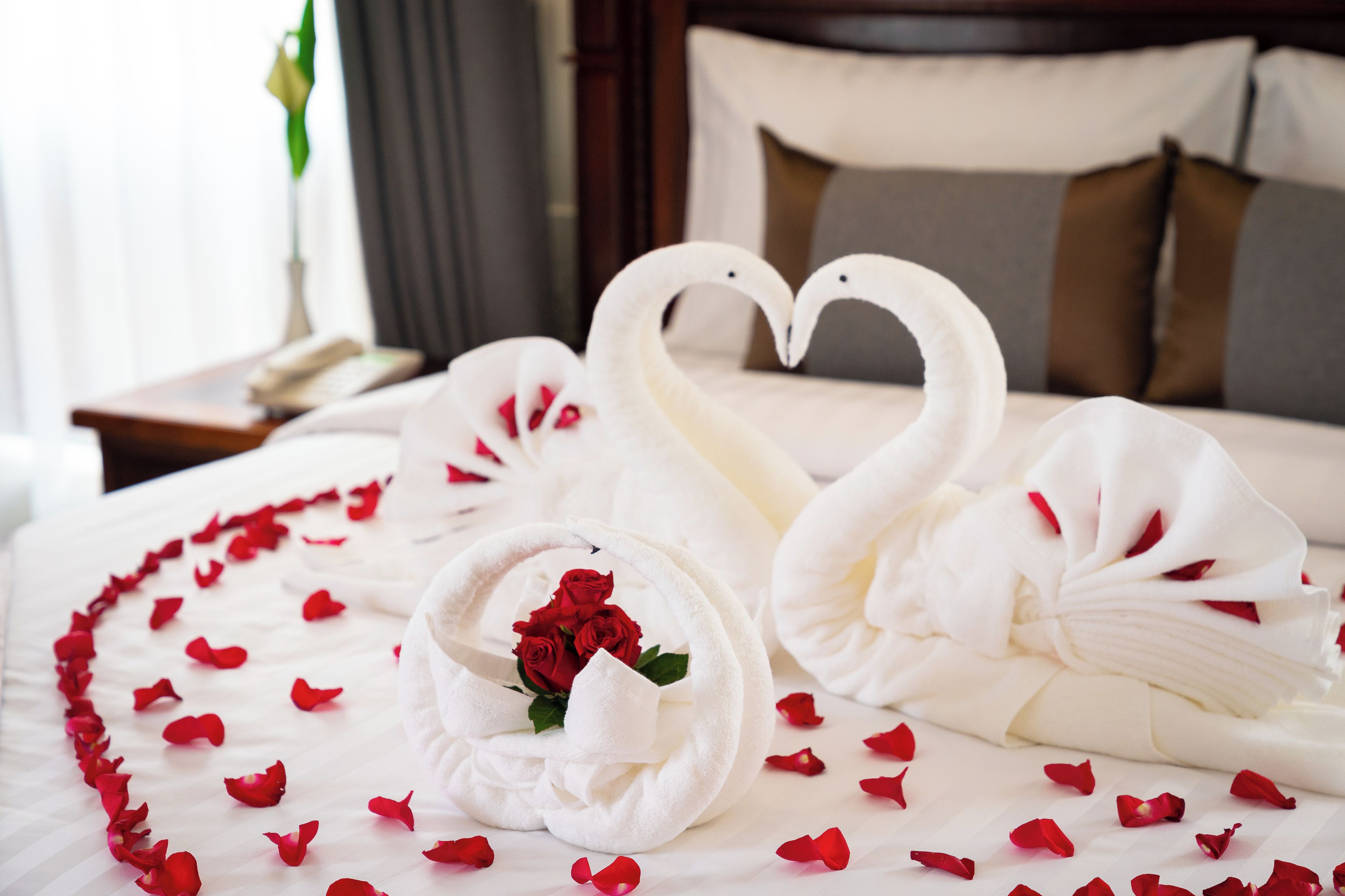 A bed with white linens, a heart-shaped arrangement of towels, and red rose petals.