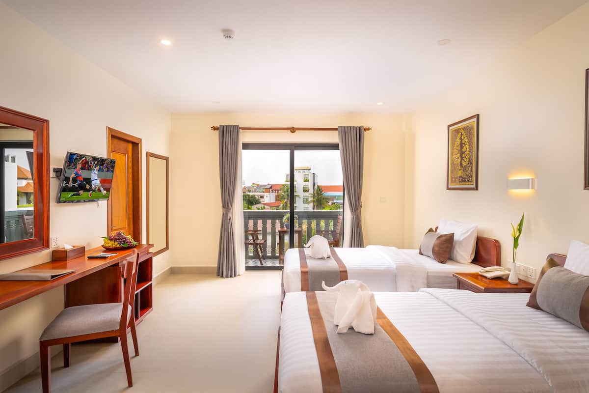 A hotel room with two beds, a desk, a chair, a TV, and a balcony with a view of buildings.