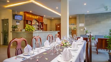 A restaurant with a long dining table set for a meal, wooden chairs, and a bar area with a television screen.