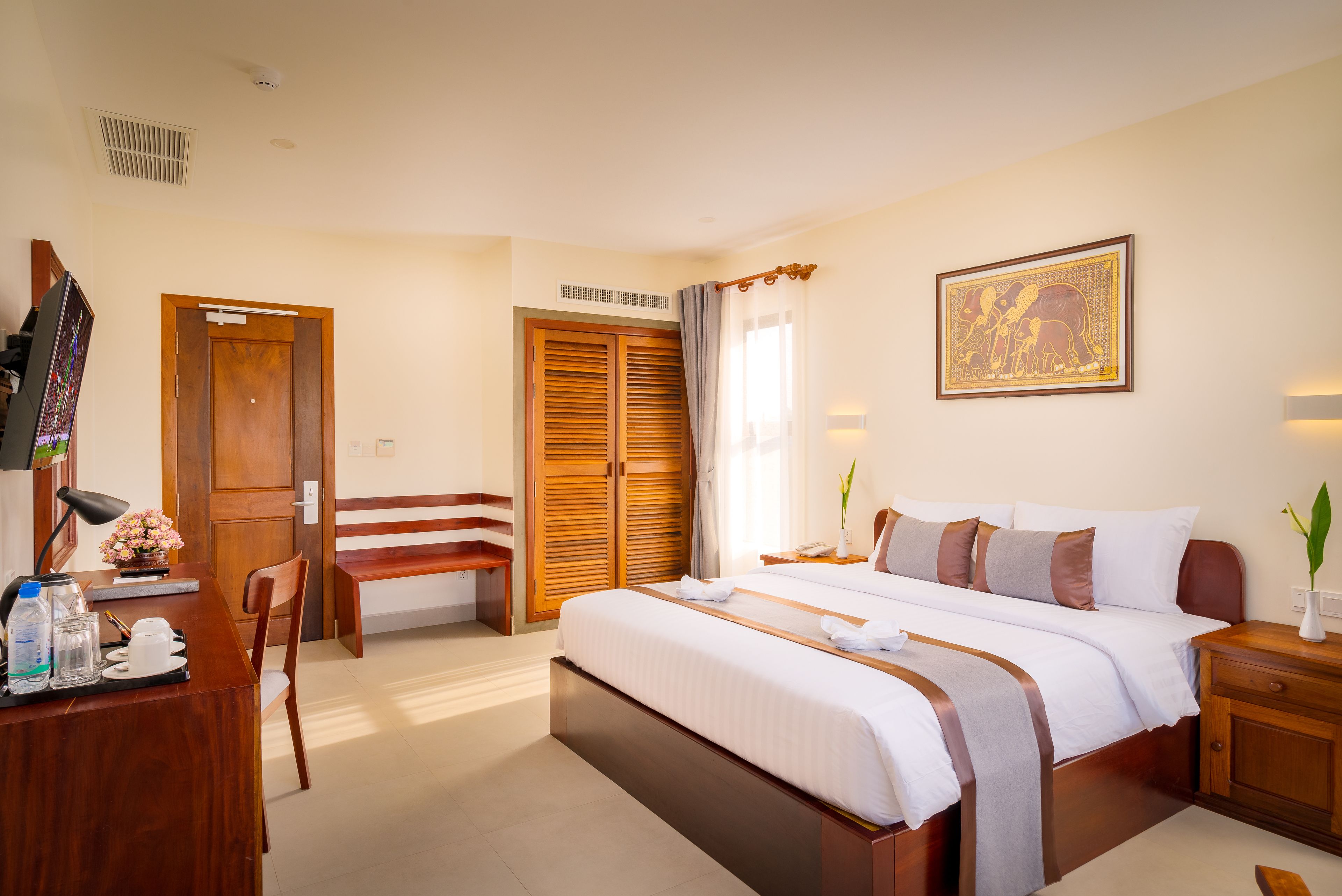 A hotel room with a large bed, wooden wardrobe, desk, and a painting on the wall.