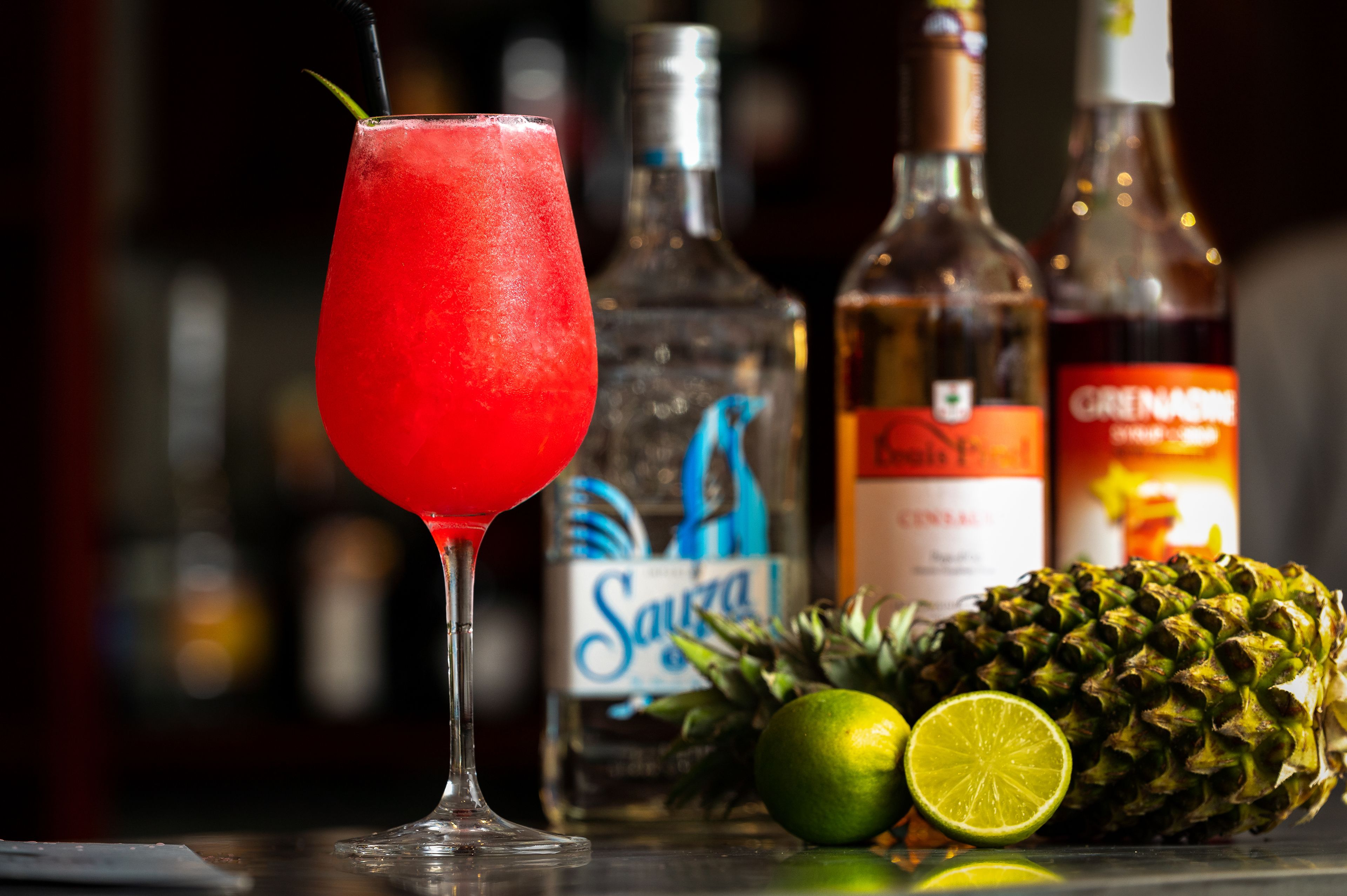 Una copa con un trago rojo, una botella de tequila Sauza, una botella de granadina, un ananá y una lima.