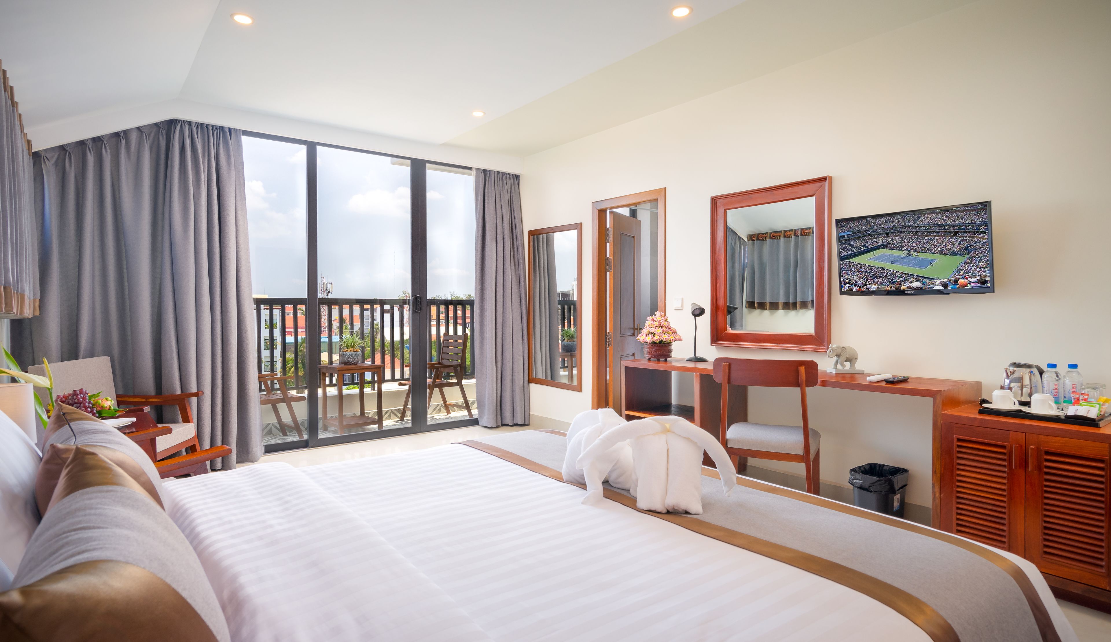 A hotel room with a large bed, a desk with a mirror, a TV, and a balcony with curtains.