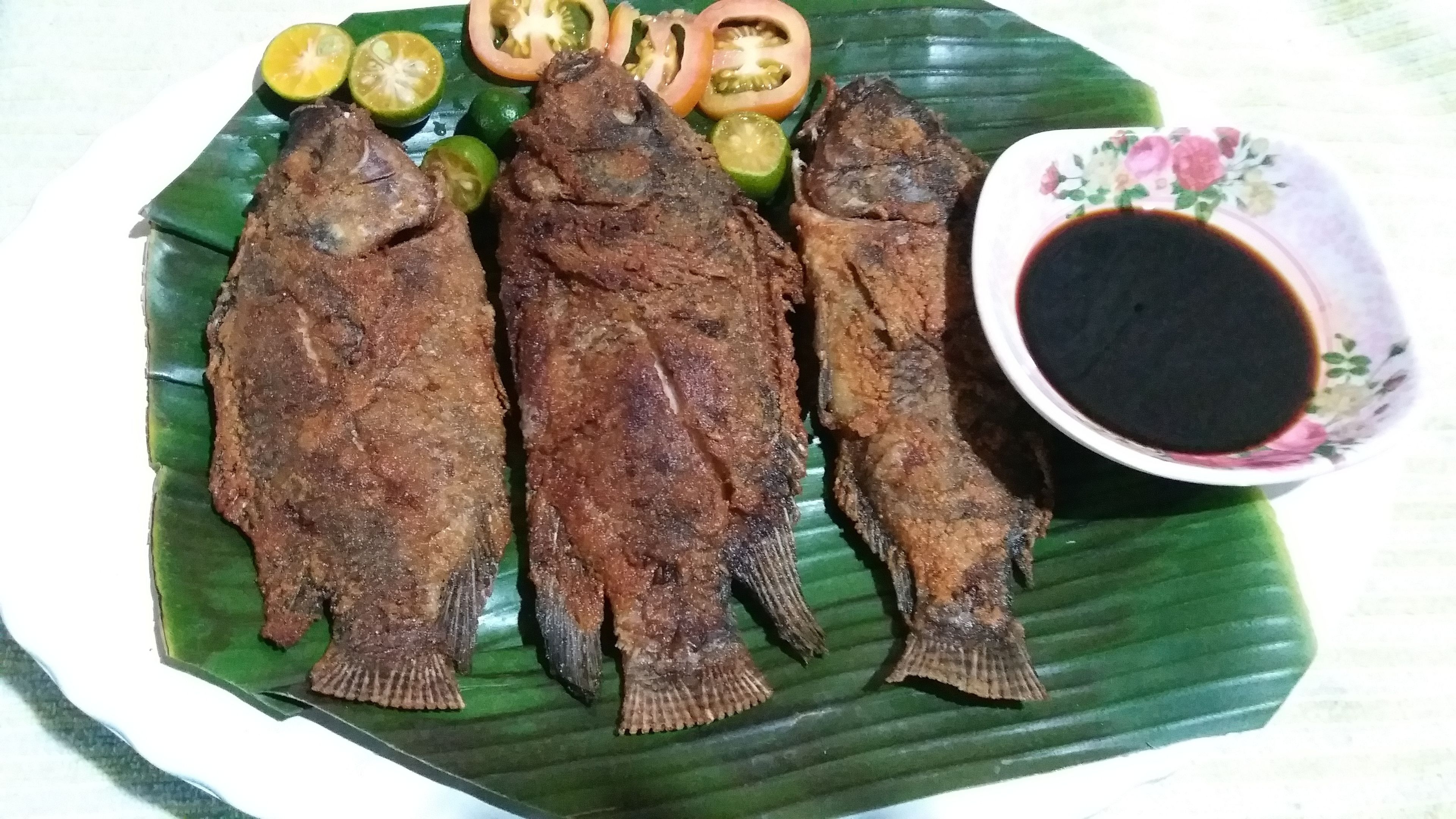 A plate with three fried fish, sliced tomatoes, and a bowl of dipping sauce.