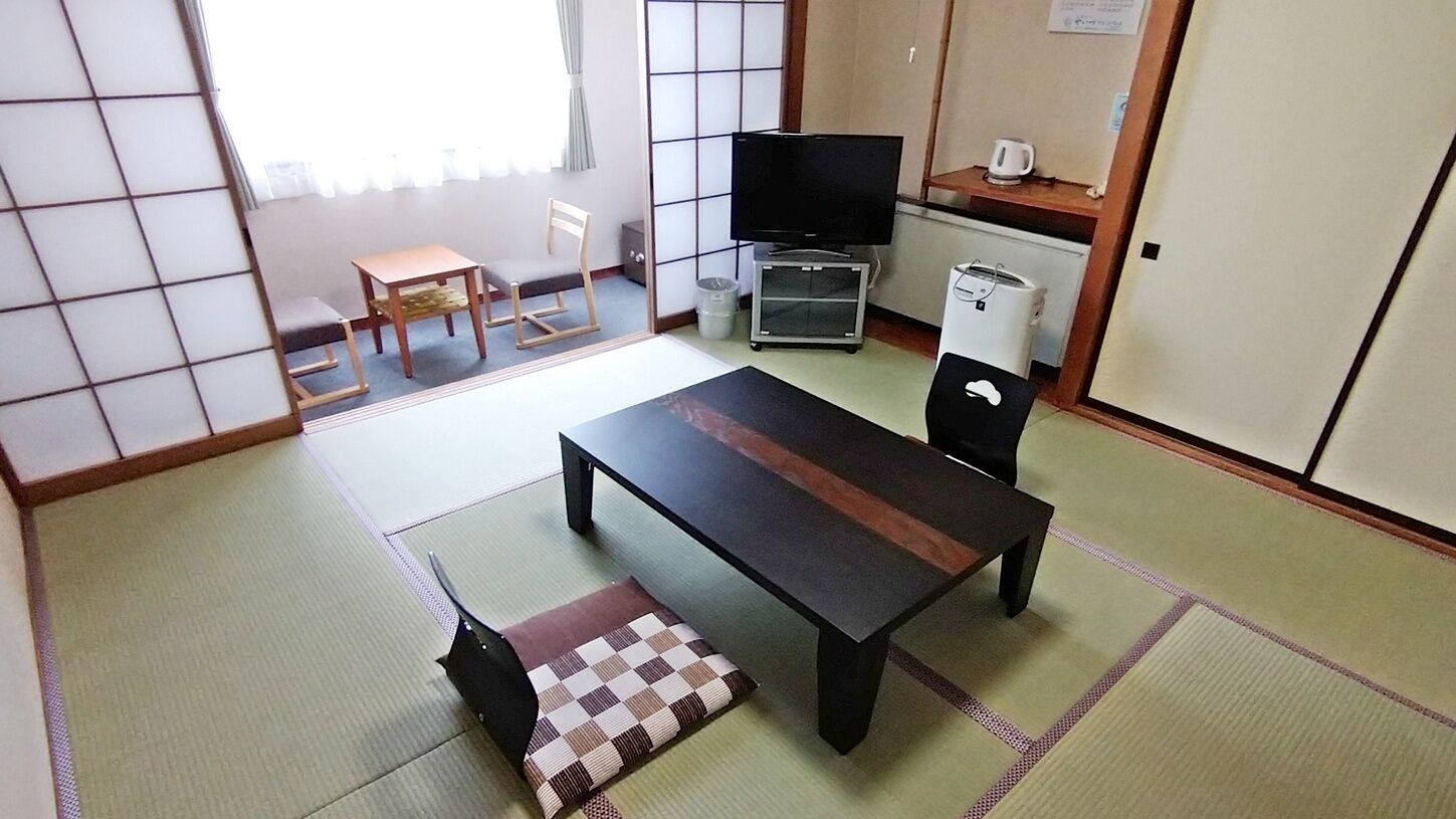 Um quarto japonês tradicional com tatames, uma mesa baixa com almofada, uma televisão e uma pequena cozinha compacta.