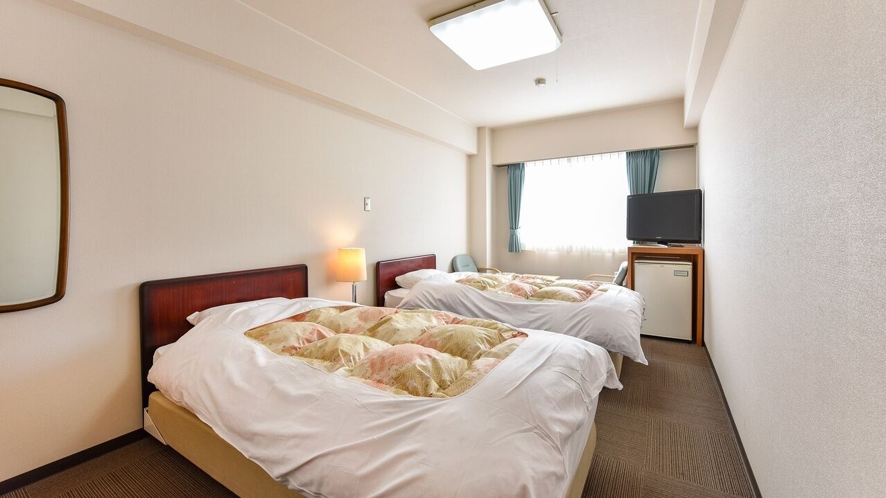 Quarto de hotel com duas camas, uma mesa de trabalho com televisão e uma janela com cortinas.