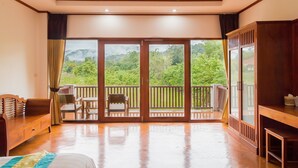 Een hotelkamer met een houten vloer, een bed, een houten kledingkast, een balkon met een tafel en stoelen, en uitzicht op groen en bergen.