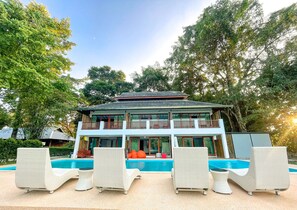 Un edificio de dos pisos con piscina, rodeado de árboles y con mobiliario exterior.
