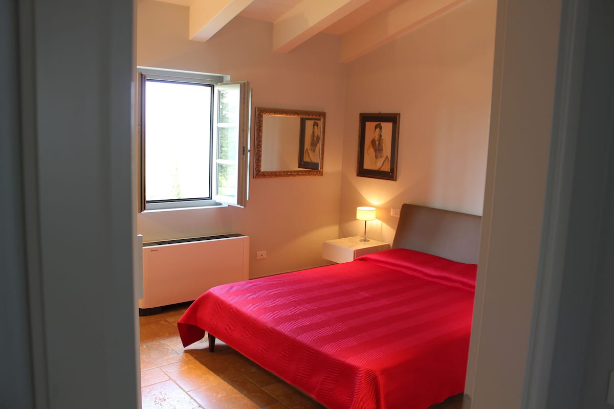 A bedroom with a bed, a window, a bedside table with a lamp, and a mirror on the wall.