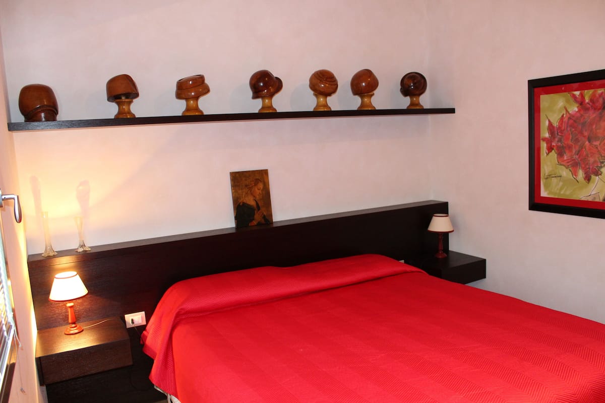 A hotel room with a red bedspread, a nightstand with a lamp, a shelf with decorative items, and a framed picture on the wall.
