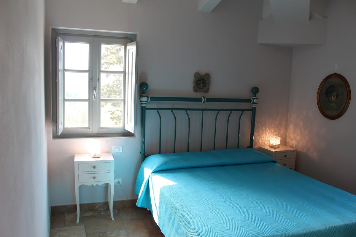 A bedroom with a bed, a nightstand, a bedside table, a window, and a wall-mounted picture.