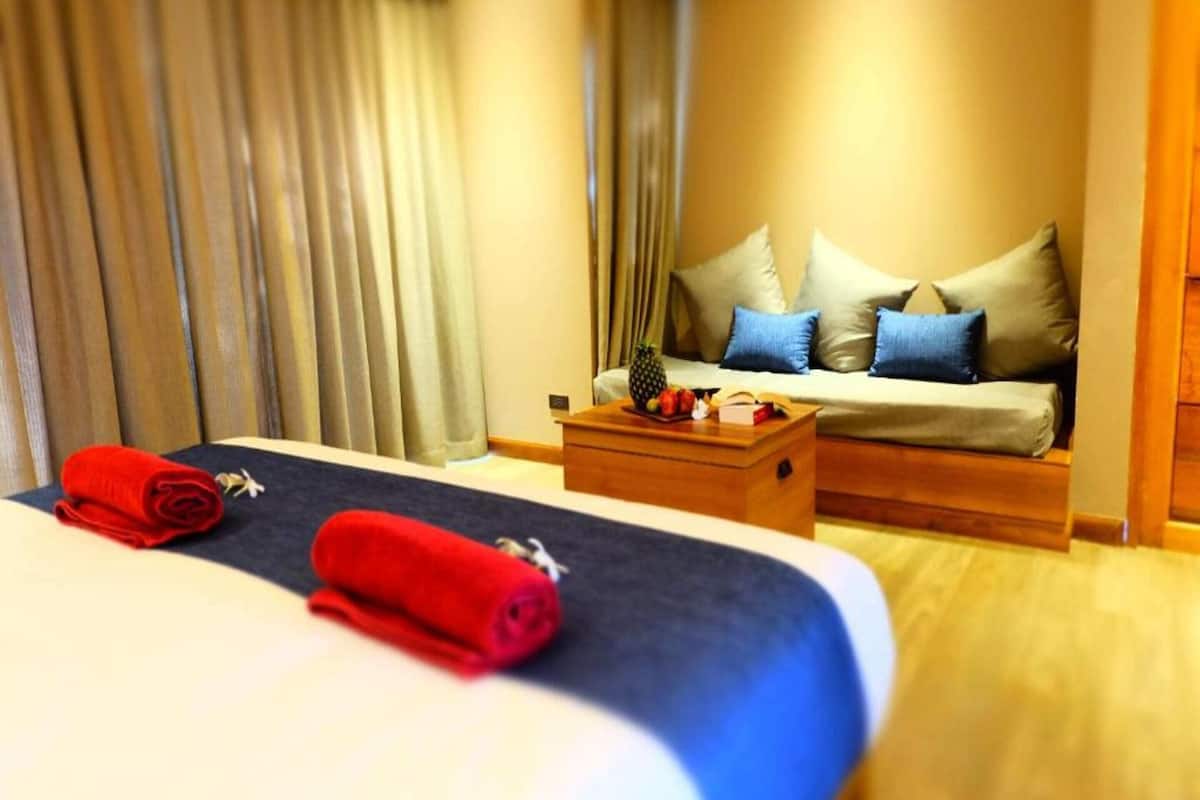 A hotel room with a bed, a wooden nightstand, a sofa with cushions, and a window with curtains.