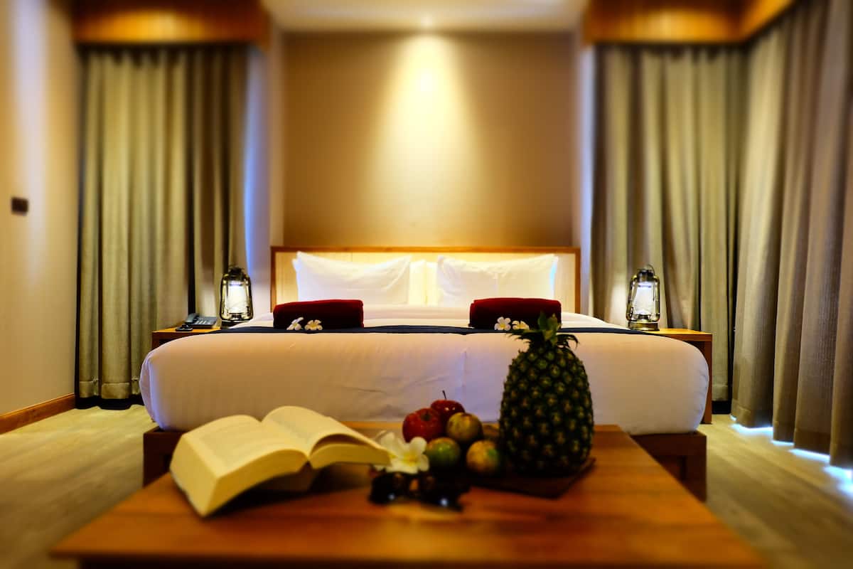 A hotel room with a large bed, two bedside tables, a nightstand, and a wooden table with fruits and an open book.