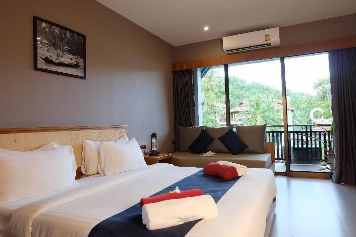 A hotel room with a large bed, a sofa, a balcony with a view, and a painting on the wall.