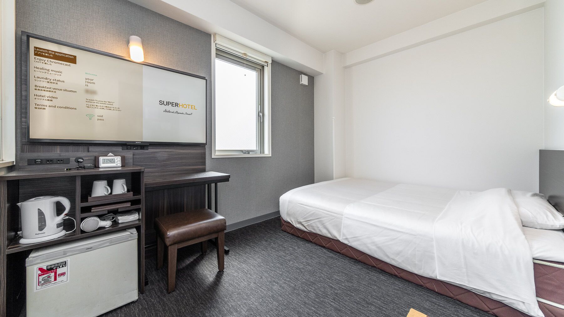 A hotel room with a bed, a desk with a coffee maker, a mini-fridge, and a TV displaying a welcome message.