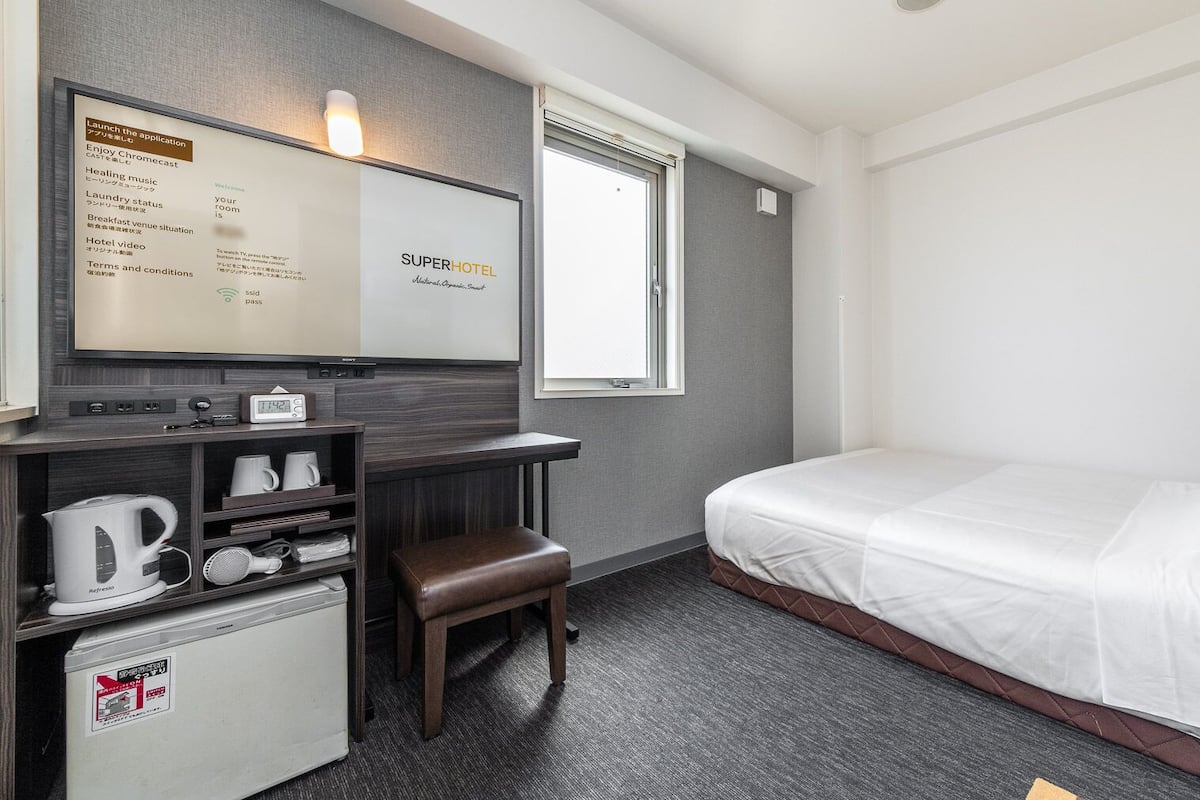 Habitación de hotel con cama, escritorio con cafetera, refrigerador pequeño y televisor mostrando un mensaje de bienvenida.