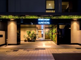 L'ingresso di un hotel con un cartello che riporta la scritta 'SUPER HOTEL'. Sono presenti piante in vaso all'esterno dell'ingresso.