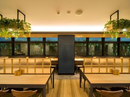 Un interior moderno con un separador negro central, mesas y sillas de madera, y plantas colgando del techo.