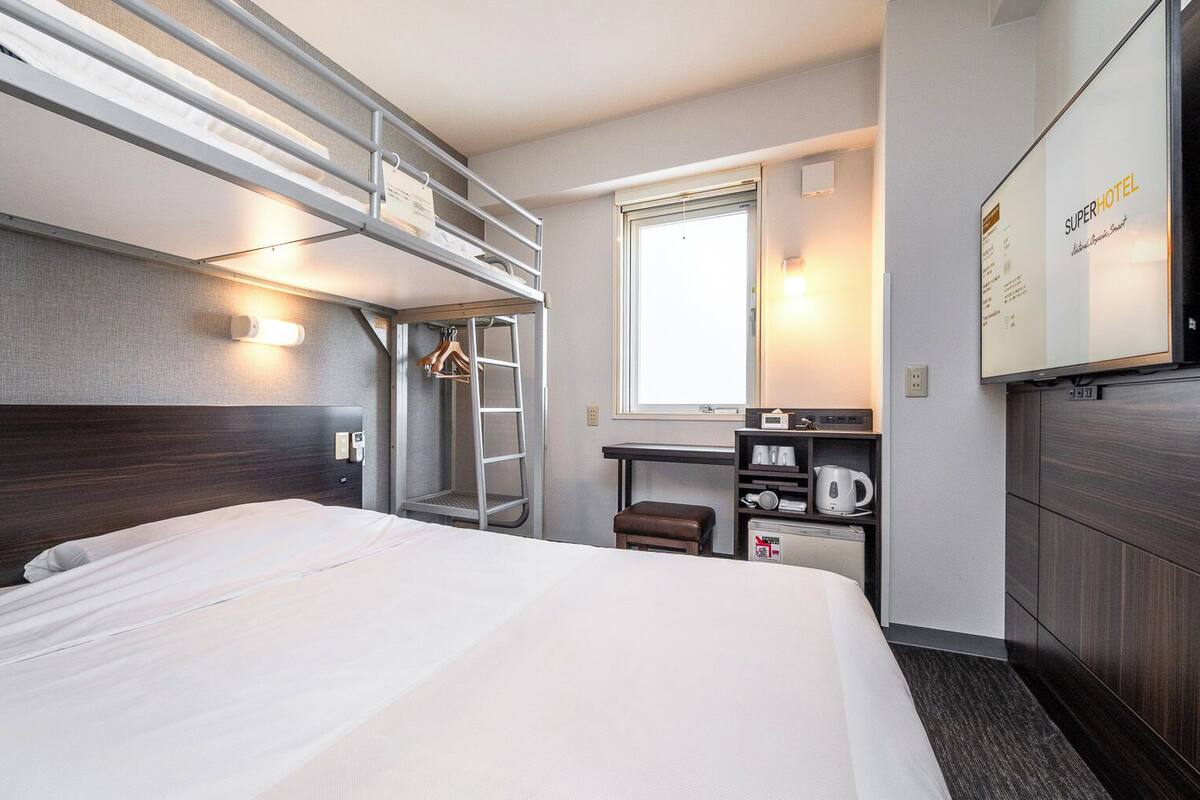 Una habitación de hotel con literas, un escritorio con hervidor, una silla y un espejo.