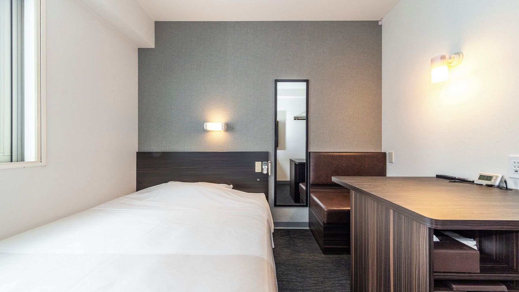 A hotel room with a bed, a desk, a mirror, and a wall-mounted light.