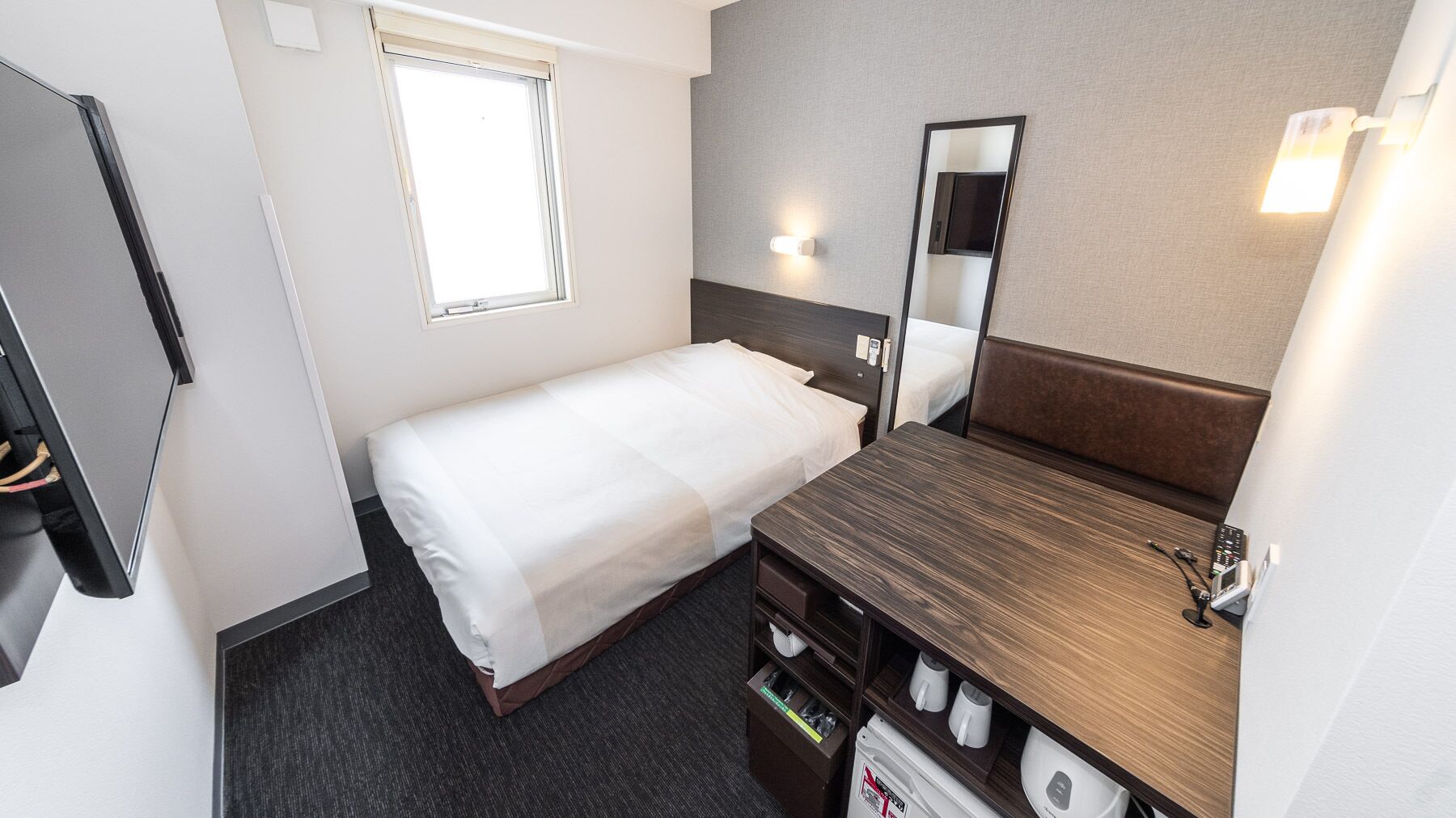 A hotel room with a bed, a wooden dresser, a mirror, a window, and a wall-mounted light fixture.