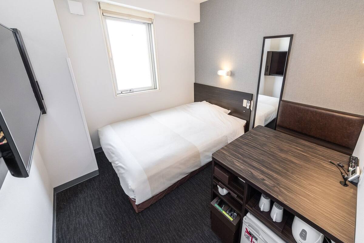 Una habitación de hotel con una cama, un aparador de madera, un espejo, un ventanal y una lámpara fijada a la pared.