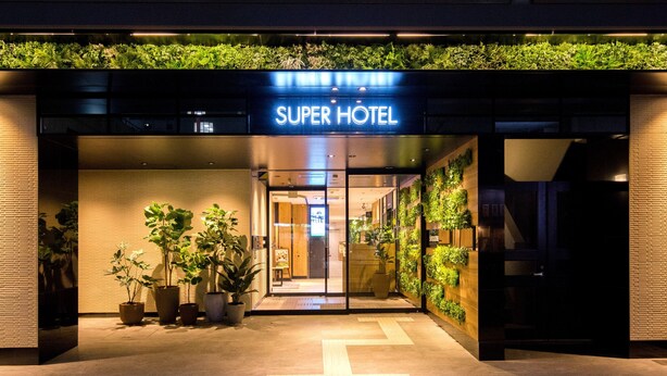 Entrada d'un hotel amb un rètol que diu 'Super Hotel', plantes en testos i un disseny modern.