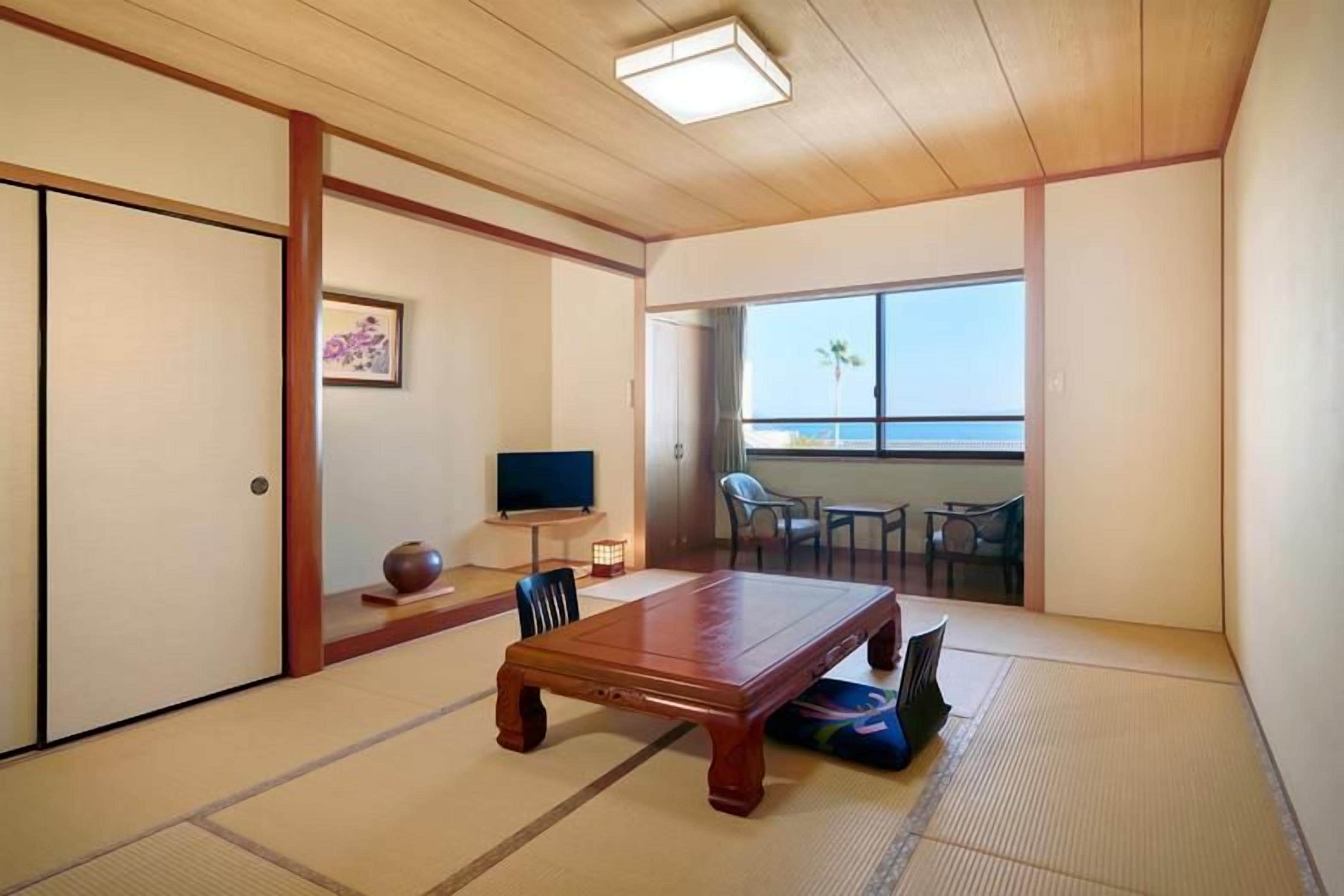 Ein Raum mit Tatami-Matten, einem Holztisch, Stühlen, einem Fernseher und Blick auf das Meer.