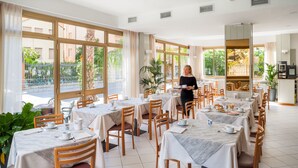 Uma ampla sala de jantar com várias mesas e cadeiras, grandes janelas com vista para o exterior, e uma área de cozinha visível ao fundo.