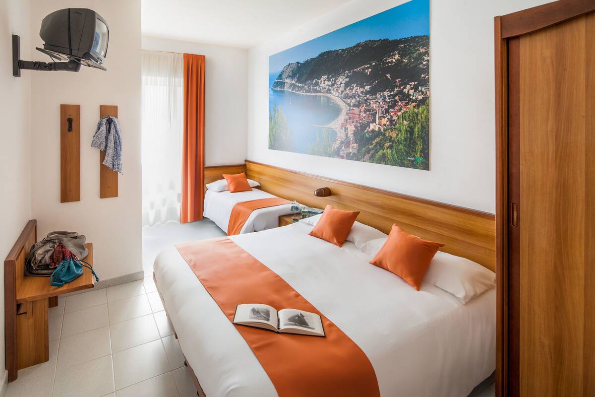 A hotel room with two beds, a wooden headboard, a large window with curtains, a TV mounted on the wall, and a painting of a coastal town.