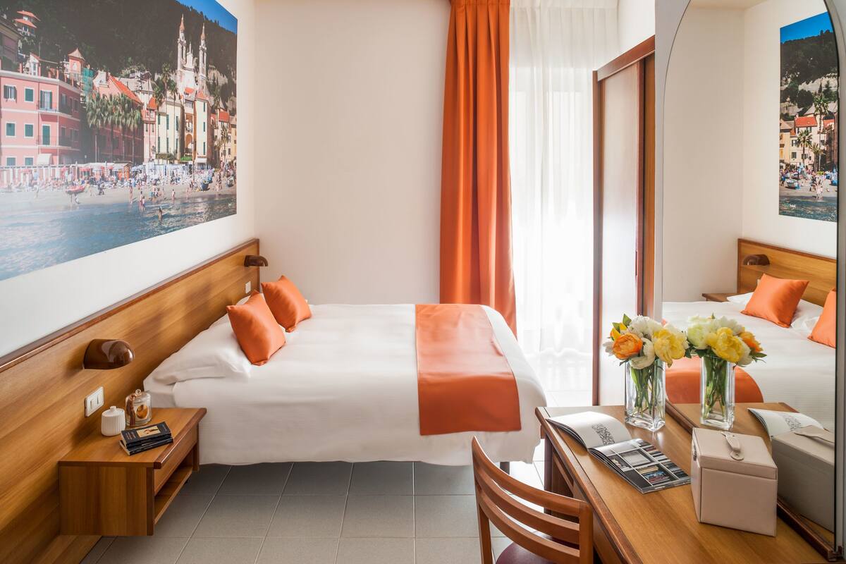 A hotel room with a bed, a desk, a chair, a nightstand, a mirror, and a large window with curtains.