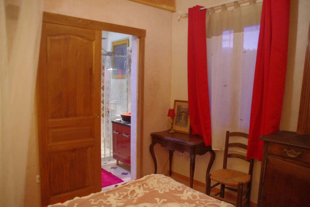 Une chambre avec une porte en bois, un lit recouvert d’une couvre-lit à motifs, une chaise en bois, une petite table sur laquelle se trouve un cadre photo et un rideau rouge.