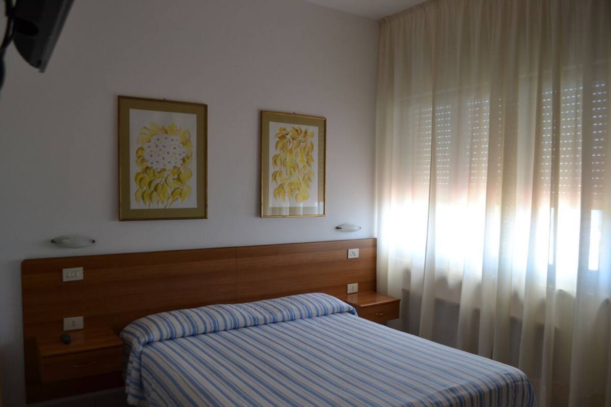 A hotel room with a wooden headboard, a bed with striped bedding, two framed floral artworks on the wall, and a window with sheer curtains.