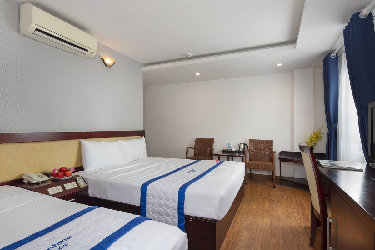 A hotel room with two beds, a desk with a chair, a small table with a kettle, and a window with curtains.