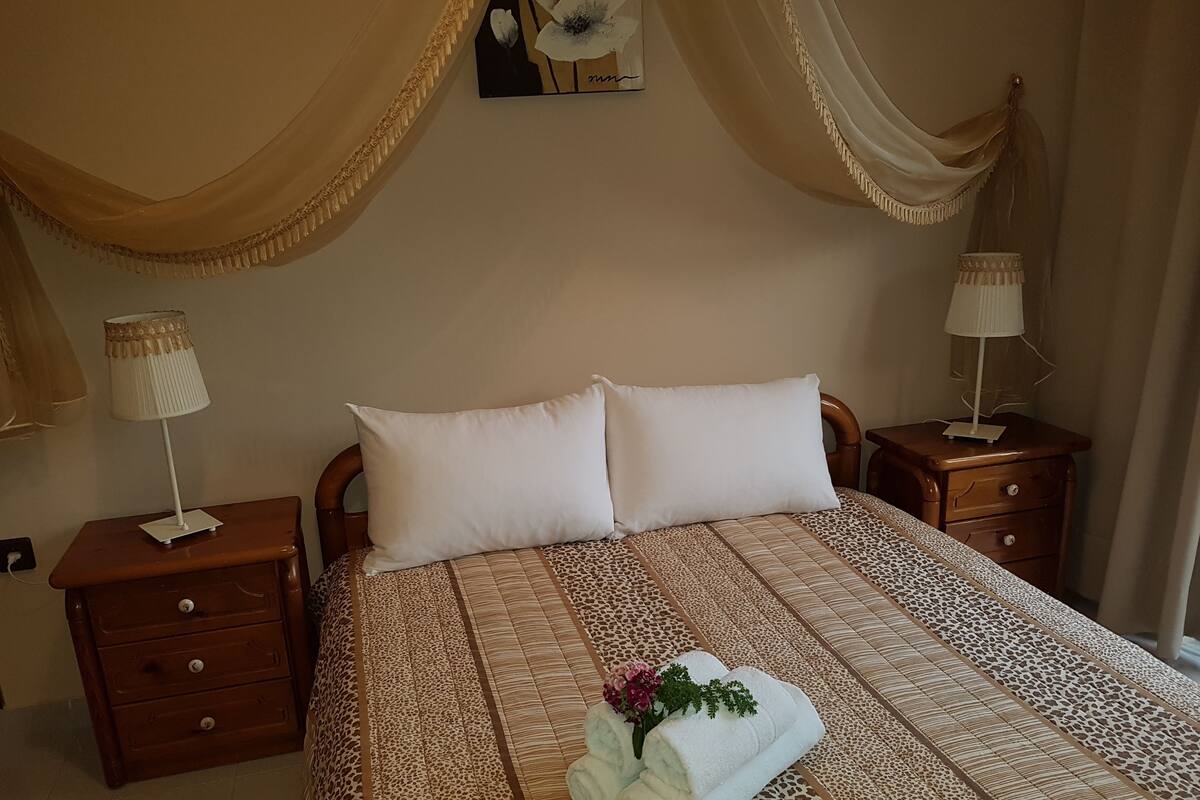 Un lit bien fait, recouvert d’une couvre-lit à motifs, avec deux oreillers et une serviette ornée de fleurs, posés sur une table de chevet en bois.