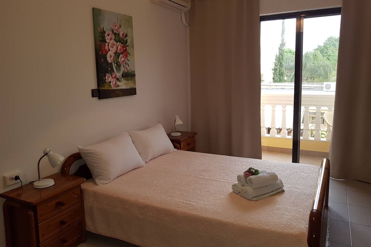 Une chambre à coucher comprenant un lit, une table de chevet, une lampe, un tableau et donnant sur le balcon extérieur.