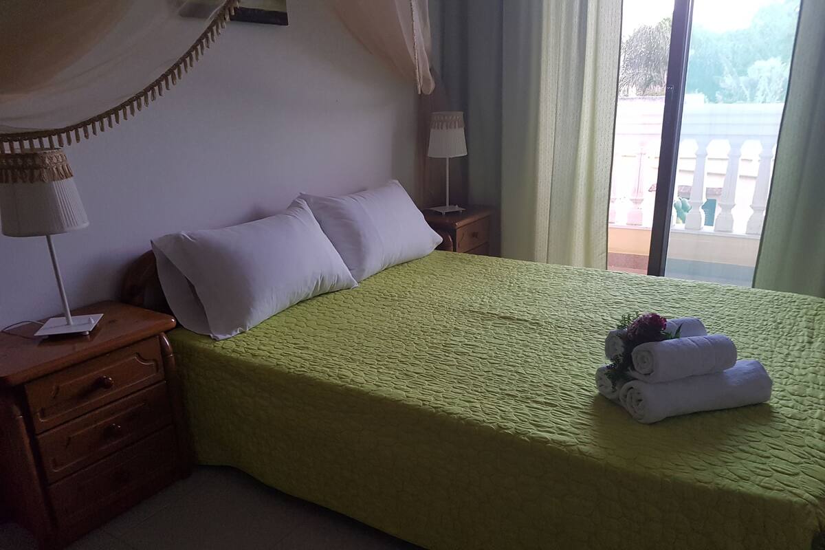 Un lit bien fait, avec une couette verte, deux oreillers et un petit bouquet de fleurs posé sur le lit.