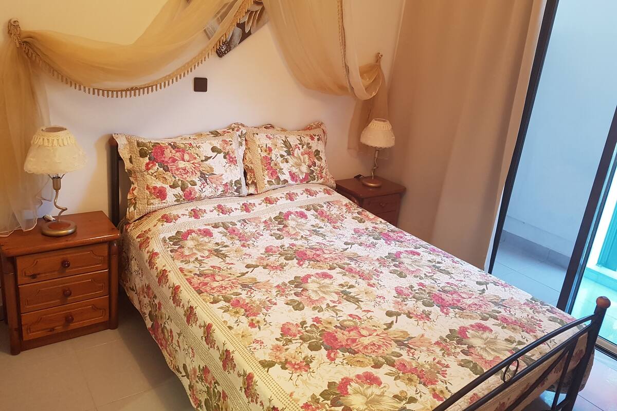 Une chambre à coucher avec une couvre-lit à motifs floraux, deux tables de chevet avec des lampes, une petite table en bois et une fenêtre avec des rideaux.