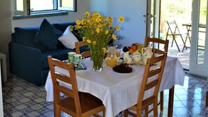 Comedor con una mesa preparada para cuatro personas, rodeado de sillas de madera, un florero con flores amarillas y un frutero.