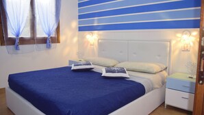 Uma cama bem arrumada com colcha azul, dois travesseiros e uma mesa de cabeceira com abajur.