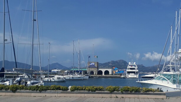 Uma marina com vários veleiros atracados, um edifício com um mural e montanhas ao fundo.