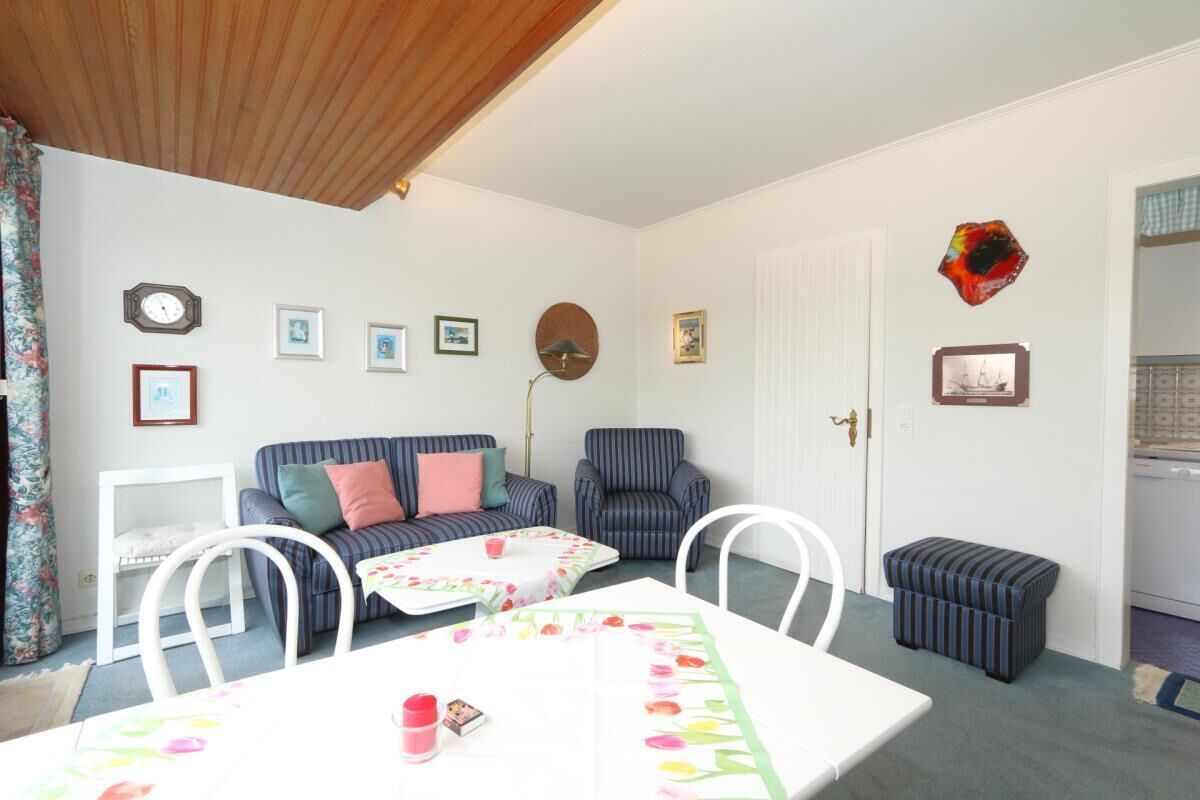 Un salon chaleureux avec une table blanche, un canapé à rayures et un fauteuil à rayures. Des tableaux encadrés ornent le mur, et une porte est agrémentée d’une plaque décorative.