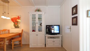 Zona pranzo con tavolo e sedie in legno, un'alzata con ante in vetro, un televisore su un mobile bianco e una porta con maniglia in ottone.
