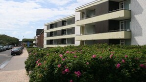 Un edificio residenziale a più piani con balconi, un'area di parcheggio e un siepe con fiori rosa.