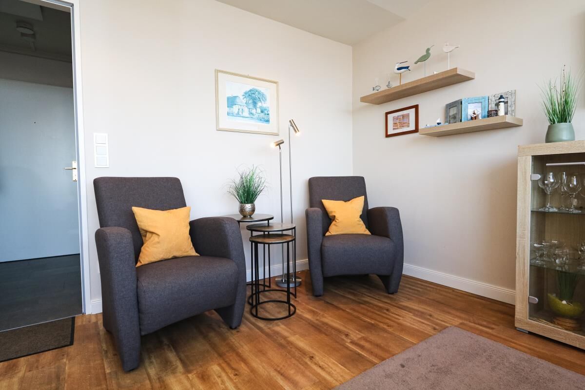 A living room with two grey armchairs, a small round table with a plant, a floor lamp, and a cabinet with glassware.