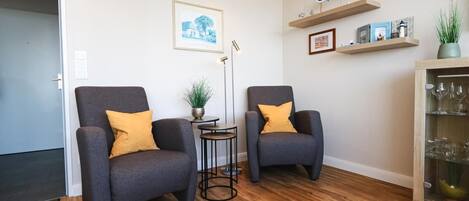 A living room with two grey armchairs, a small round table with a plant, a floor lamp, and a cabinet with glassware.