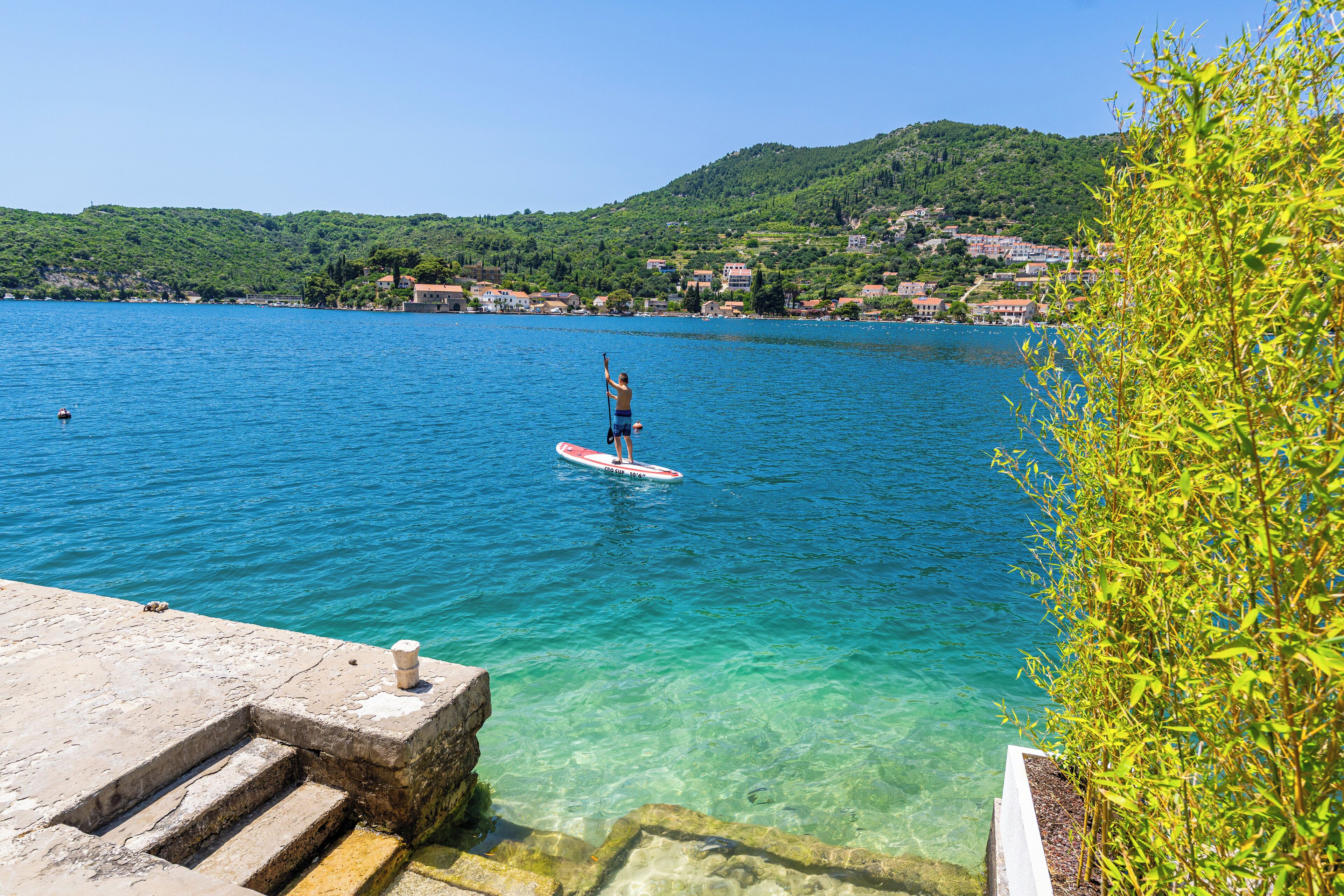 Una vista despejada de un lago con una persona haciendo paddleboard, una escalera de piedra que desciende al agua y una colina con edificios al fondo.