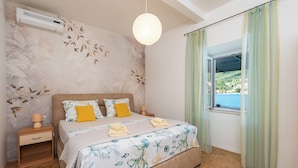 A bedroom with a bed, nightstands, an air conditioner, a hanging light, a window with curtains, and a view of a body of water. - Apartment Cecilija - waterfront with sunbeds (Zaton)
