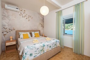 A bedroom with a bed, nightstands, an air conditioner, a hanging light, a window with curtains, and a view of a body of water.